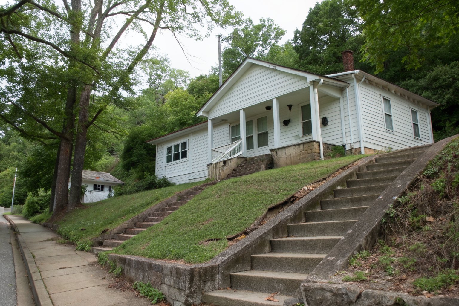 White wood-sided bungalow high on a wooded hillside, with long concrete steps up from the street